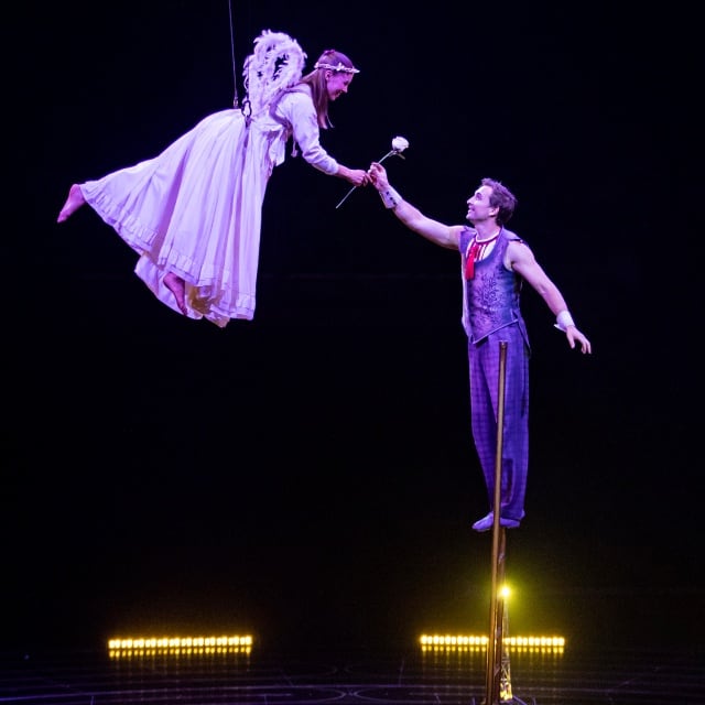 A female acrobat wearing an angel costume is hanging from the ceilling as a male performer hands her a flower.